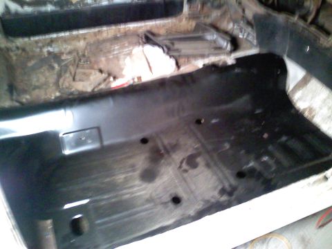 1969 Ford Mustang - The interior floor pan of a vehicle, showing a fresh black coating and pre-drilled holes for mounting components.