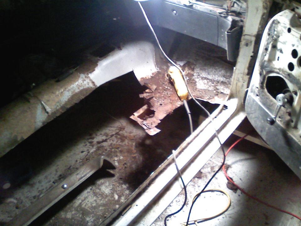 Interior of a car showing significant rust on the floor pan and a yellow tool. Door panel is removed