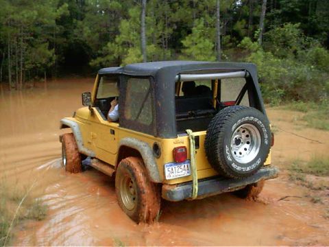 1985 Ford Bronco - A yellow Jeep Wrangler navigates through a muddy, water-filled trail surrounded by trees.