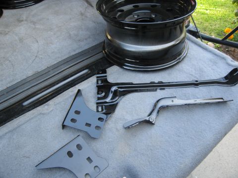 1986 Ford Bronco - Various black painted automotive parts, including a steel wheel and metal brackets, are laid out on a gray surface.