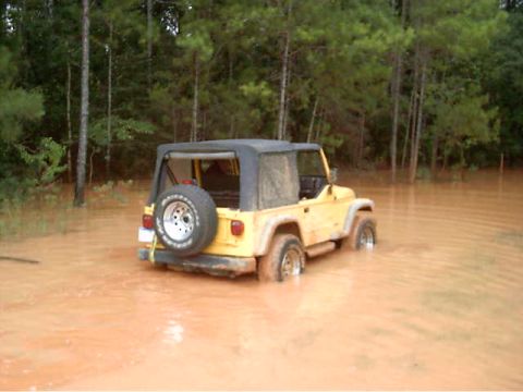 1985 Ford Bronco - A yellow Jeep Wrangler is partially submerged in muddy water, surrounded by a wooded area.