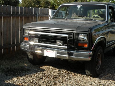1986 Ford Bronco - This Ford Bronco features a custom grille and bull bar, parked in a gravel driveway next to a wooden fence.