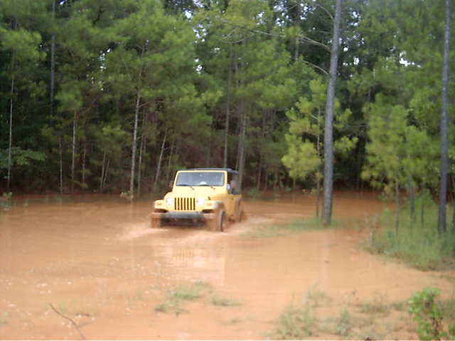Yellow Jeep Wrangler driving through deep muddy orange water on a wooded trail