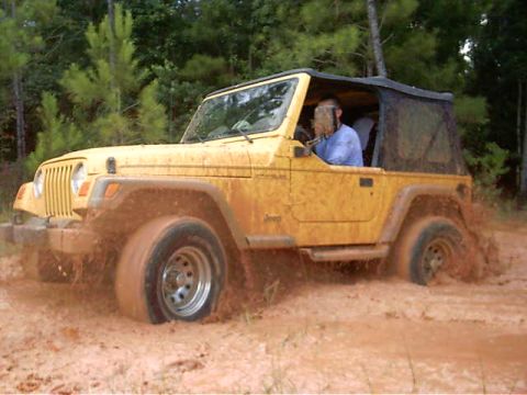 1985 Ford Bronco - A yellow Jeep Wrangler navigates through a muddy terrain, kicking up a spray of dirt.