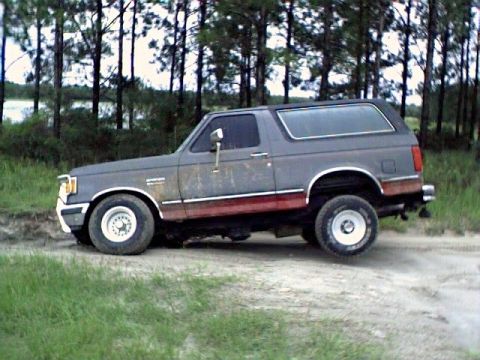 1991 Ford Bronco - Driver-side profile of a two-tone gray-over-red 1987-91 Ford Bronco with hardtop, chrome west-coast mirrors, white steel wheels and mud-spl…
