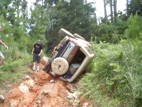 1985 Ford Bronco - An overturned SUV rests precariously in a muddy ditch, surrounded by dense foliage and trees.