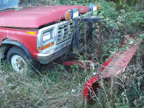 1988 Ford F-150 - This red truck, likely from the 1980s, is partially obscured by vegetation and fitted with a front plow.