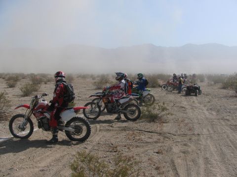 1996 Ford C-350 (Centurion Conversions) - A group of dirt bike riders and an ATV are gathered in a dusty desert area, possibly before or after an event.