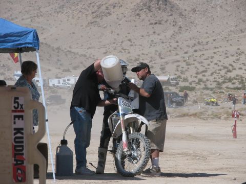 1996 Ford C-350 (Centurion Conversions) - A dirt bike is being refueled by two men under a blue tent at an outdoor event with campers and vehicles in the background.