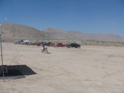1996 Ford C-350 (Centurion Conversions) - A dirt bike rider navigates a dusty track with parked vehicles and tents in the distance under a clear blue sky.