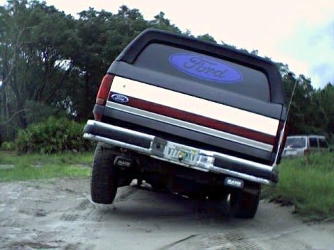 1991 Ford Bronco - A Ford Bronco is shown from the rear on a dirt path with trees and another vehicle in the distance.