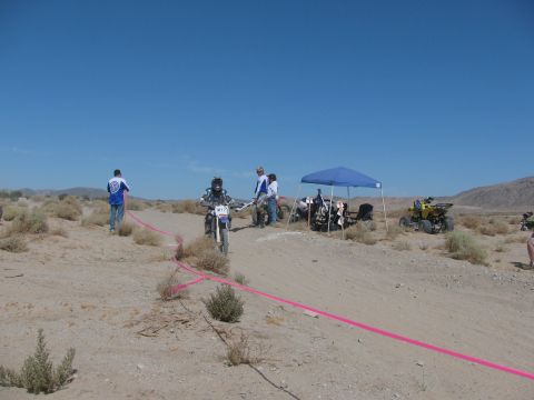 1996 Ford C-350 (Centurion Conversions) - A dirt bike rider numbered M17 navigates a desert course, with ATVs and spectators gathered under a blue canopy in the background.