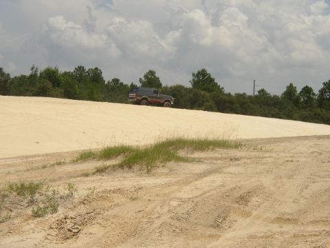 1991 Ford Bronco - A gray Ford Bronco with a red lower stripe crests the top of a large white sand hill at an off-road pit, seen small in the distance from th…