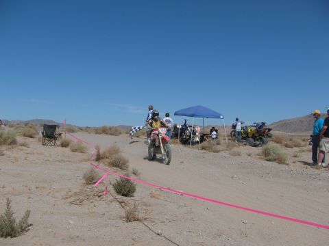1996 Ford C-350 (Centurion Conversions) - A dirt bike crosses the finish line at a desert race event with spectators and ATVs present.