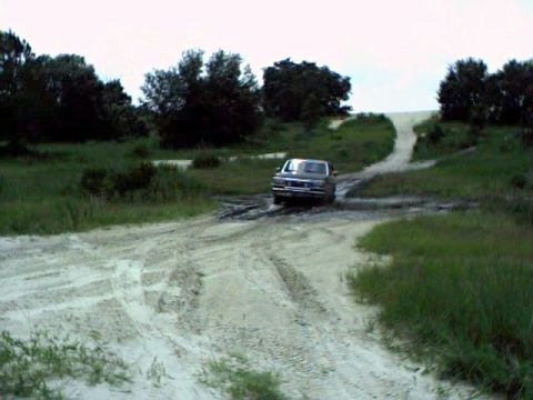 1991 Ford Bronco - Ford pickup truck navigating a muddy dirt road with grassy hills and trees.