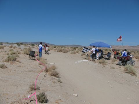 1996 Ford C-350 (Centurion Conversions) - A desert race event is underway with participants on dirt bikes and ATVs, marked by checkered flags and tents.