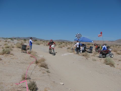 1996 Ford C-350 (Centurion Conversions) - A desert race event with a rider on a dirt bike approaching a checkpoint with spectators and an ATV in the background.
