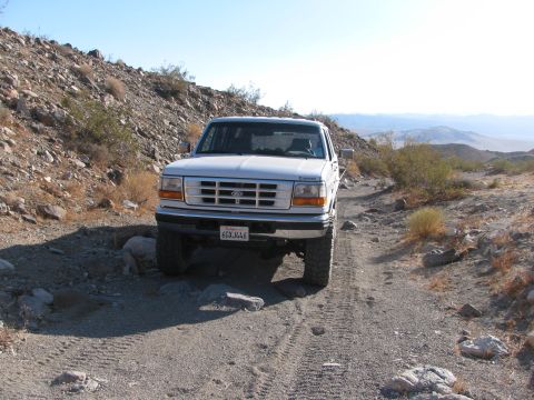 1996 Ford C-350 (Centurion Conversions) - This white Ford F-Series truck navigates a rocky dirt road in a desert landscape.