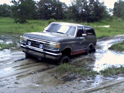 1991 Ford Bronco - A gray two-tone Ford Bronco sits axle-deep in a wet, rutted mud pit in an open grassy field, shown from the front driver-side corner. The t…