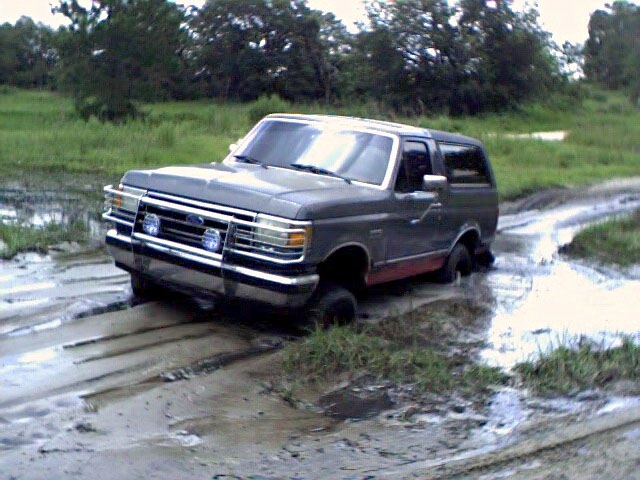 Gray Ford Bronco stuck in a rutted water-filled mud pit in a field, chrome brush guard on front