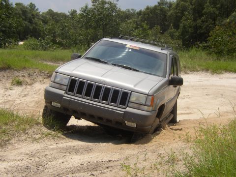 1991 Ford Bronco - Jeep Grand Cherokee stuck in sand on a dirt track with trees in the background.