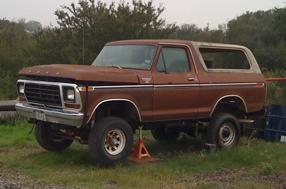 Lifted brown Ford Bronco with white top and rusty hood, parked on grass with a jack stand under one side