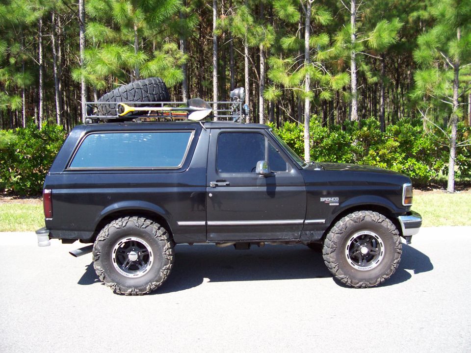 Side view of a black 1994 Ford Bronco with a roof rack carrying a spare tire and shovel