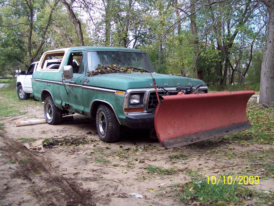 Green Ford Bronco with a red snow plow attached, parked outside. Another white vehicle is behind it