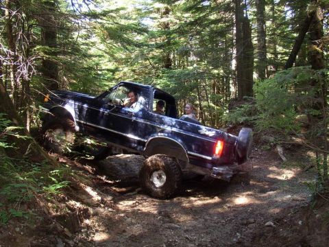 1988 Ford Bronco - A dark blue Ford Bronco navigates a challenging off-road trail through a dense forest.