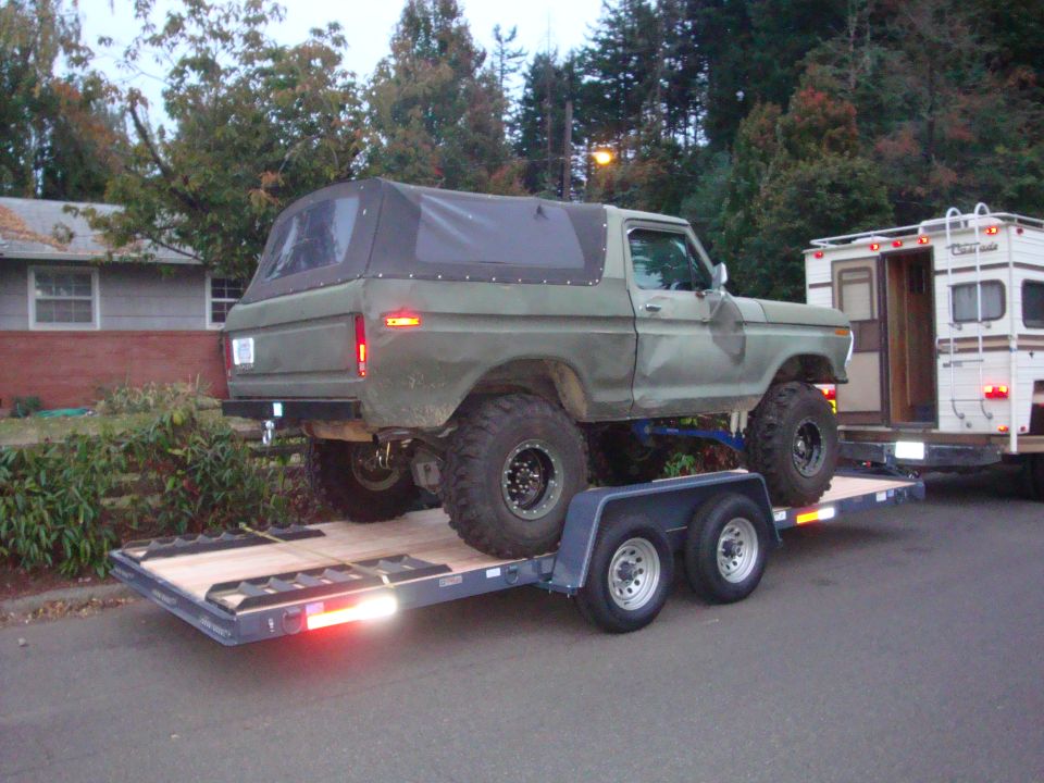 Olive green Ford Bronco with a black soft top, lifted suspension, and large off-road tires, loaded onto a dual-axle trailer