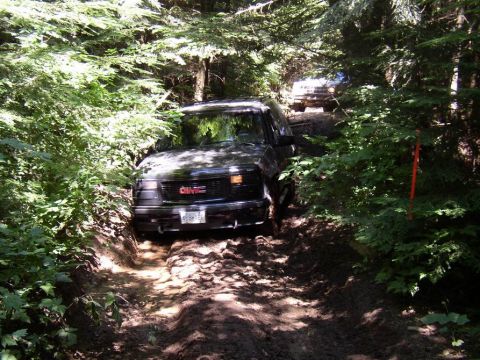 1988 Ford Bronco - GMC SUV navigating a challenging muddy off-road trail surrounded by dense trees.