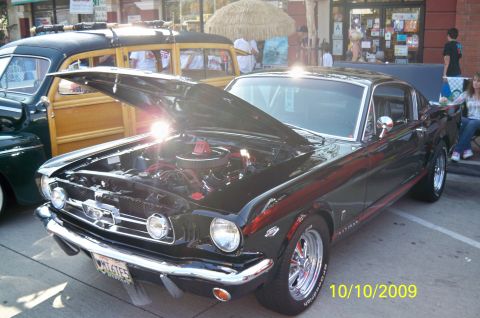 1985 Ford Bronco - A black 1965 Ford Mustang Fastback is displayed with its hood open, parked beside a vintage woodie wagon.