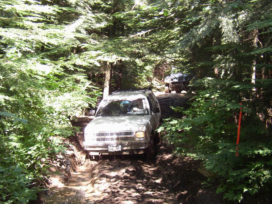 Two SUVs driving on a muddy, wooded trail. The lead vehicle is light-colored and appears to be struggling on the terrain