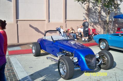 1985 Ford Bronco - A blue hot rod is parked on a street with people sitting in chairs nearby. A second blue car is visible to the right.