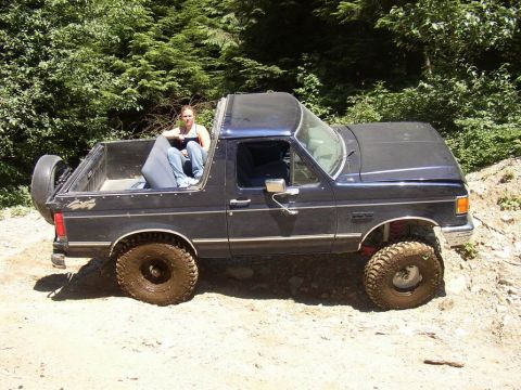 1988 Ford Bronco - This dark blue Ford Bronco, likely a 1987 model, is equipped with off-road tires and has a woman sitting in its open bed.