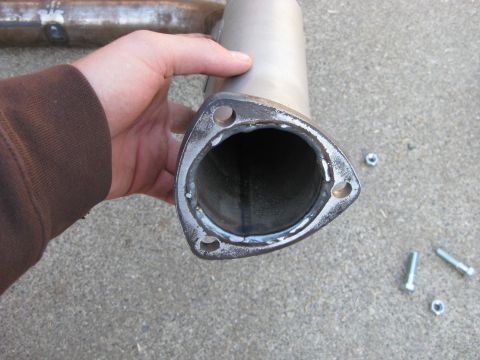 1995 Ford Bronco - A hand in a brown sleeve holds up the end of an exhaust pipe fitted with a freshly welded three-bolt triangular collector flange, with loos…