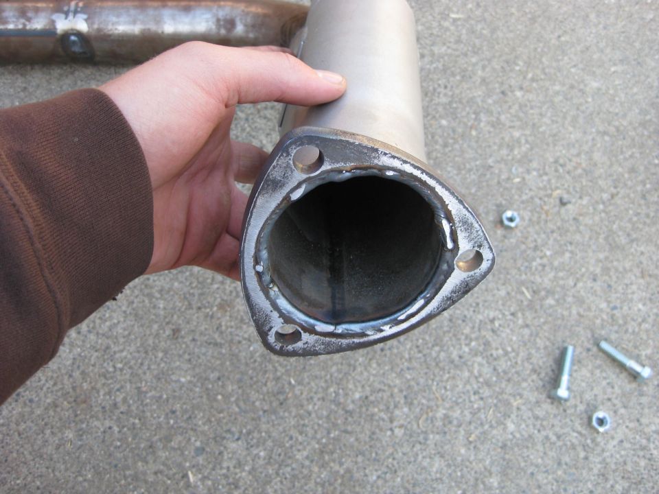 Hand holding an exhaust pipe with a newly welded three-bolt flange, bolts on concrete