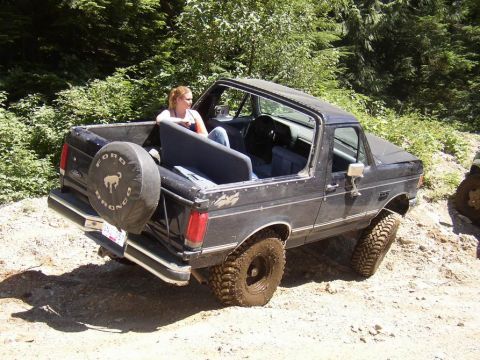1988 Ford Bronco - Black Ford Bronco with spare tire on back, driving off-road. Woman sits in the open rear.
