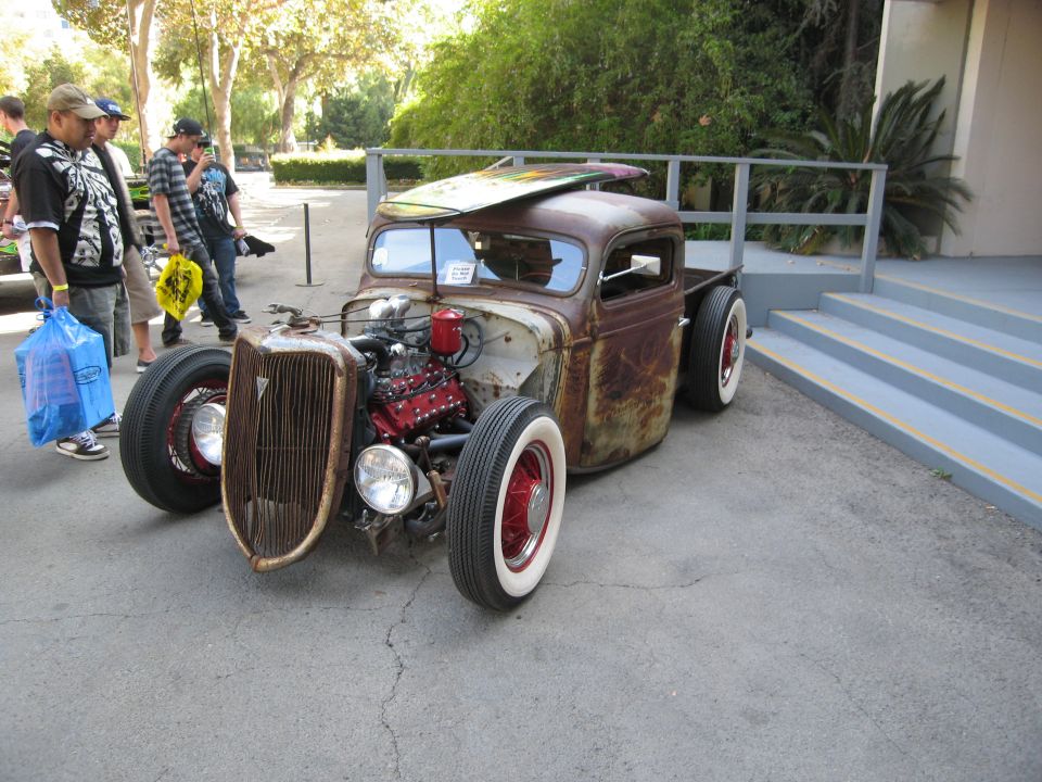 Rusty rat rod pickup truck with exposed engine and surfboard on roof