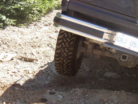 1988 Ford Bronco - The rear of a dark SUV is shown on a rough, gravelly surface with vegetation in the background.