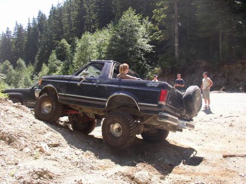 1988 Ford Bronco - A dark blue 1987 Ford Bronco navigates an off-road trail, showing its rear profile and spare tire mounted externally.