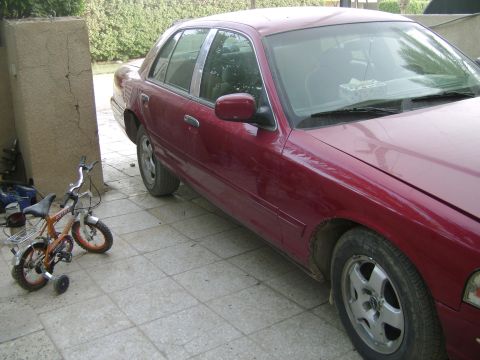 1998 Ford Crown Victoria - A maroon 1998 Ford Crown Victoria is parked on a tiled driveway next to a small bicycle.