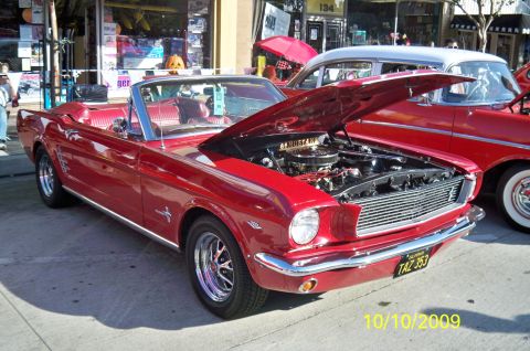 1985 Ford Bronco - A red 1965 Ford Mustang convertible is displayed with its hood open, revealing the engine, next to another red vehicle.