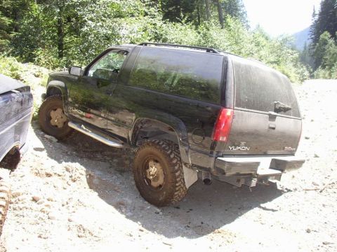 1988 Ford Bronco - A black GMC Yukon SUV is parked on a dirt road with trees in the background.