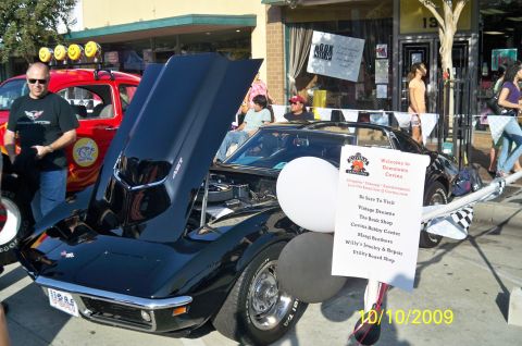 1985 Ford Bronco - A black Corvette is displayed with its hood open at an outdoor event, with a red vehicle and onlookers nearby.