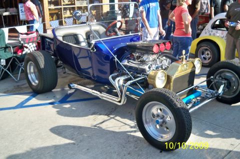 1985 Ford Bronco - A custom blue hot rod with a prominent V8 engine and chrome exhaust is displayed at an outdoor event, with other vehicles and people in the…