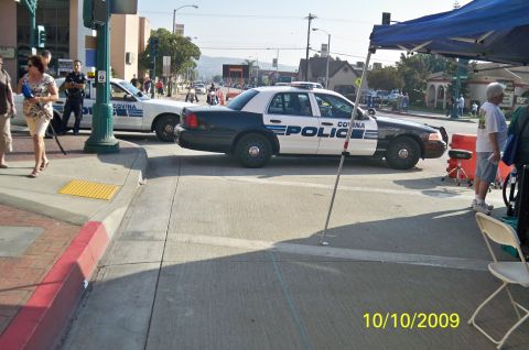 1985 Ford Bronco - A black and white police car is parked on the street, with people and other vehicles visible in the background.