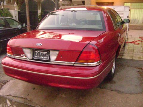 1998 Ford Crown Victoria - This red Ford sedan is parked outside, with another vehicle visible to the left.