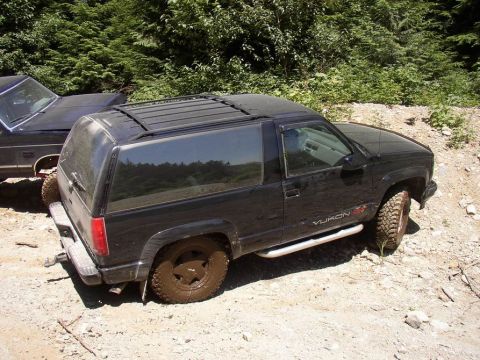 1988 Ford Bronco - A black GMC Yukon GT sits off-road, its tires caked in mud, beside another dark vehicle.