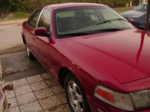 1998 Ford Crown Victoria - This red 1998 Ford Crown Victoria is parked on a tiled surface next to a building.